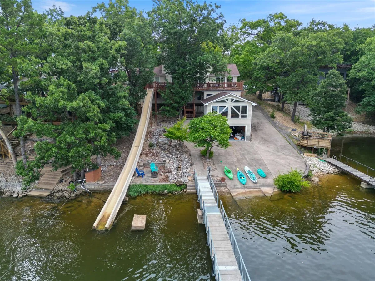 A vibrant exterior scene showing the large engineered water slide curving into the lake, flanked by a spacious dock, kayaks stacked at the ready, and lush greenery surrounding the area. The house with expansive windows sits in the background, overlooking the activity and the water.