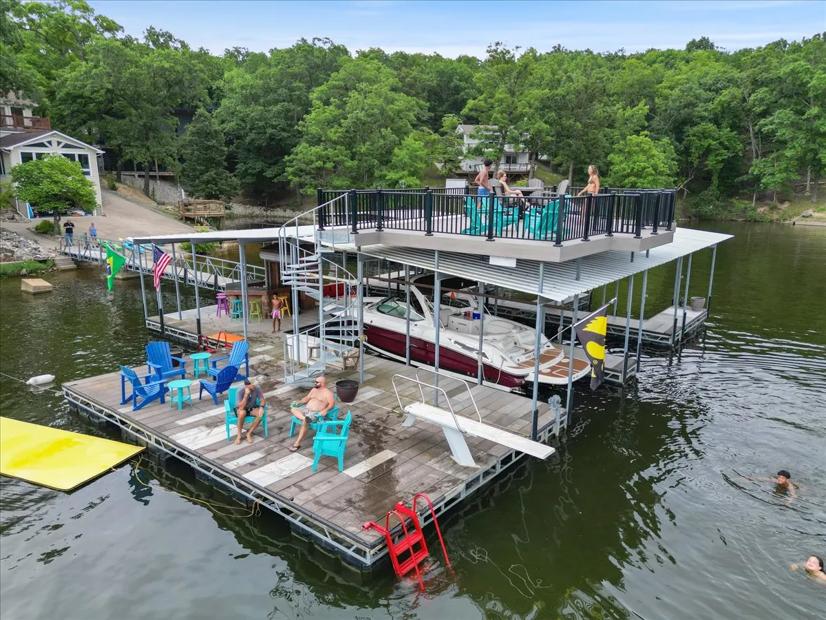The image shows a bustling floating dock with colorful chairs, swimmers in the water, a boat moored nearby, and people socializing as the sun sets. There’s a festive, communal atmosphere of summer fun on the water.
