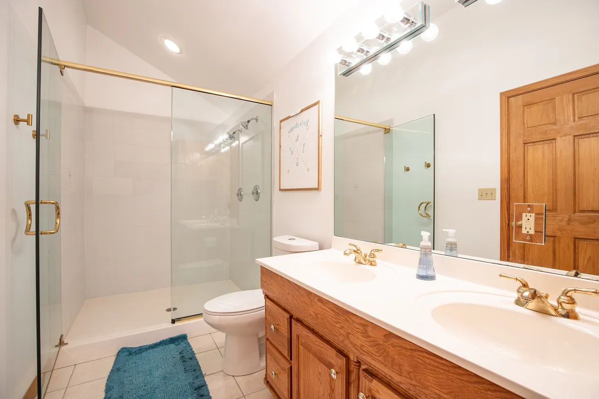 A spacious bathroom with a double sink vanity and a sleek glass shower enclosure, accented by framed artwork, all bathed in calm, natural light. The scene is tidy and serene, evoking a sense of luxury and convenience.