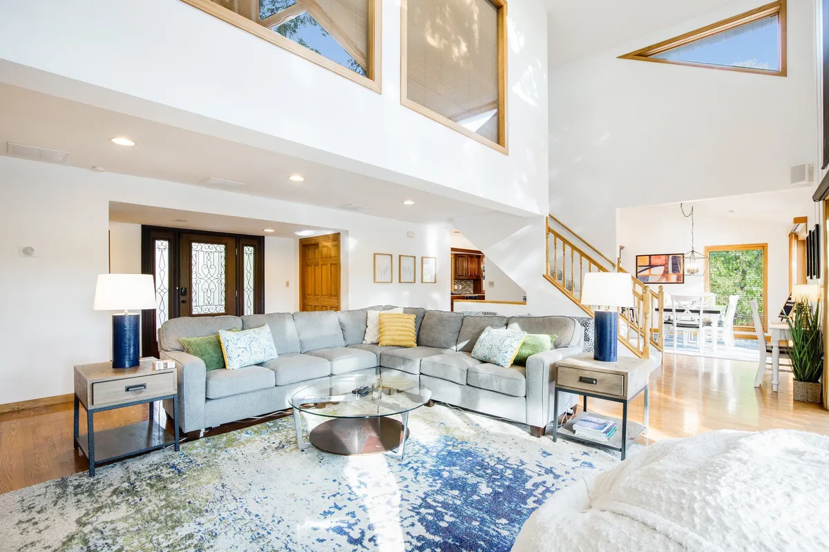 A large sectional sofa sits in a cozy, well-lit space, with a glass coffee table, decorative rug, and expansive windows. This room is designed for relaxation, socializing, and family gatherings, with natural light creating a cheerful and inviting mood.