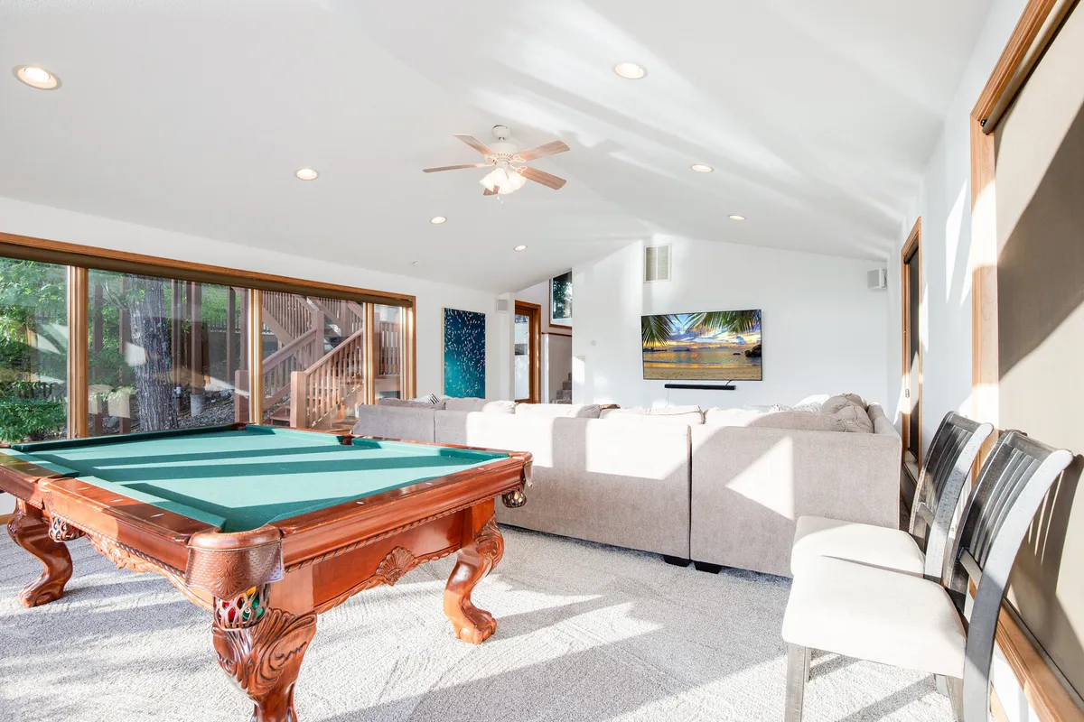 A well-lit interior scene with a pool table, large windows letting in natural light, comfortable seating, and a television. The setup encourages casual gatherings and fun, with plenty of room for both play and relaxation.