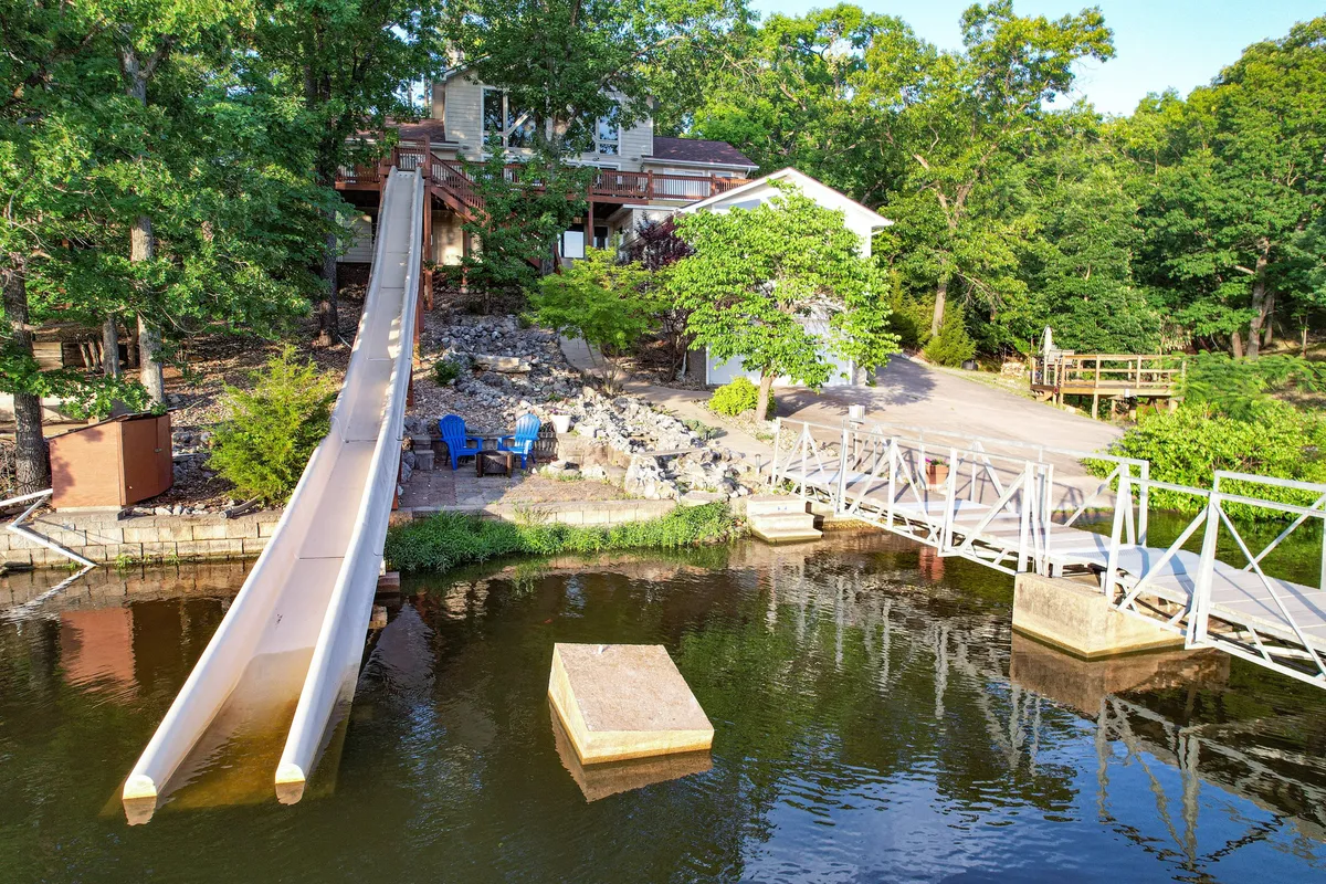 This lively exterior scene showcases a dock with a water slide, lounge chairs, and a landscaped garden by the water's edge. The area is set up for swimming, sunbathing, and family gatherings, with activities designed for both kids and adults to enjoy the lakeside setting.