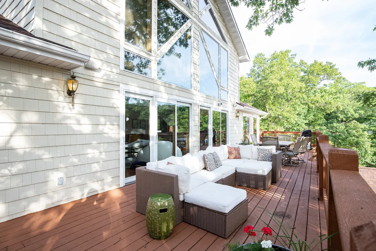 This exterior image features a spacious deck area with comfortable seating surrounded by lush greenery. Large windows allow natural light to stream into the house, suggesting an easy flow between indoor and outdoor living. The setting is serene and welcoming, ideal for gatherings and relaxing while enjoying views of the natural surroundings.