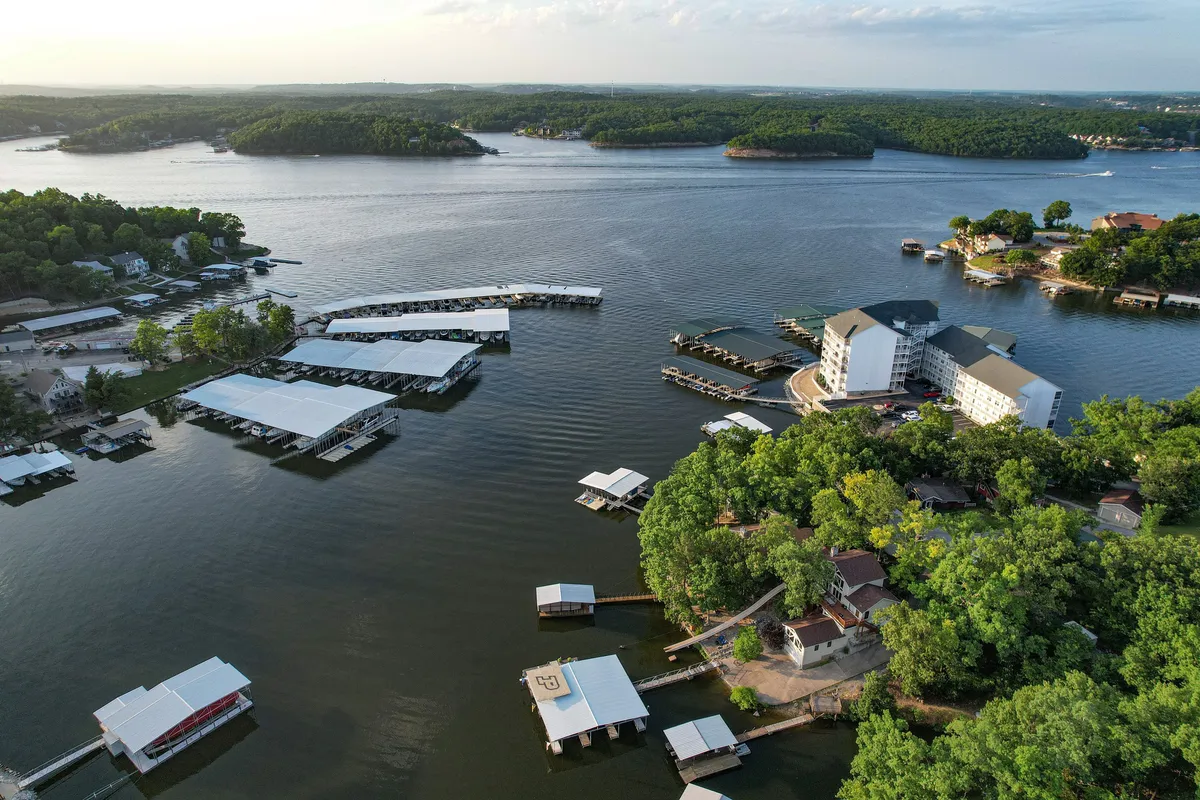 An elevated view of Lake of the Ozarks with marinas, boats, lush greenery, and buildings lining the shoreline. The calm water and natural surroundings suggest a wide range of outdoor activities and the vibrant lakeside community.