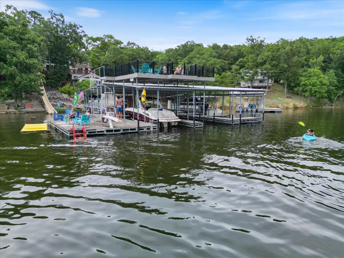 A lively dock scene featuring the water slide, a kayaker gliding by, and several people socializing and sunbathing on the dock. Boats are moored nearby, and the atmosphere is casual and joyful, with the lake shimmering under the sun.