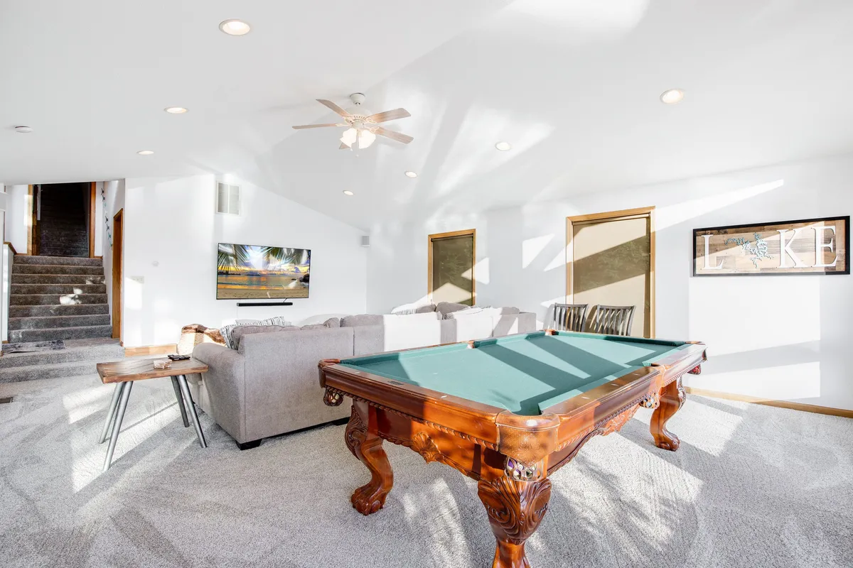 A vibrant interior space featuring a billiards table in the foreground, a comfortable sofa, a television, and stairs leading up. The room is designed for socializing, playing pool, watching TV, and relaxing with friends or family.