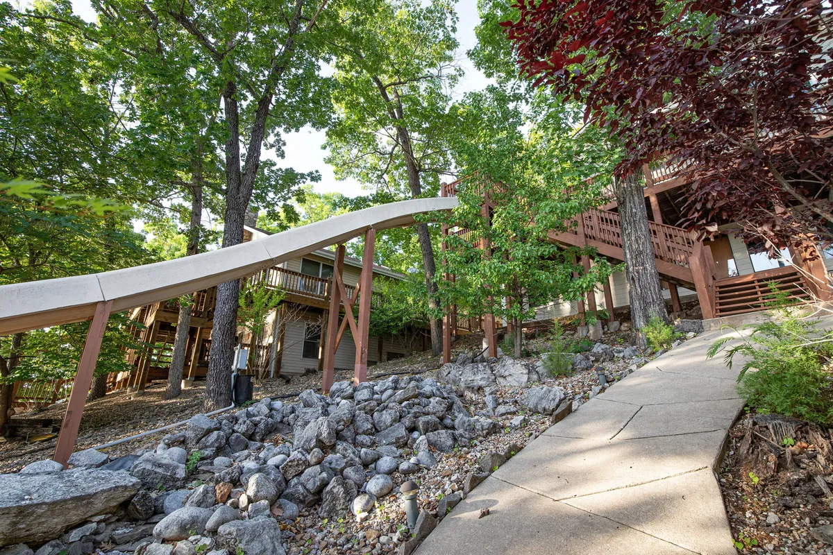 A playful outdoor scene featuring a slide integrated into stone landscaping, trees, a deck, and a winding pathway. The space is designed for both family fun and relaxation, with a clear sense of adventure and natural beauty.