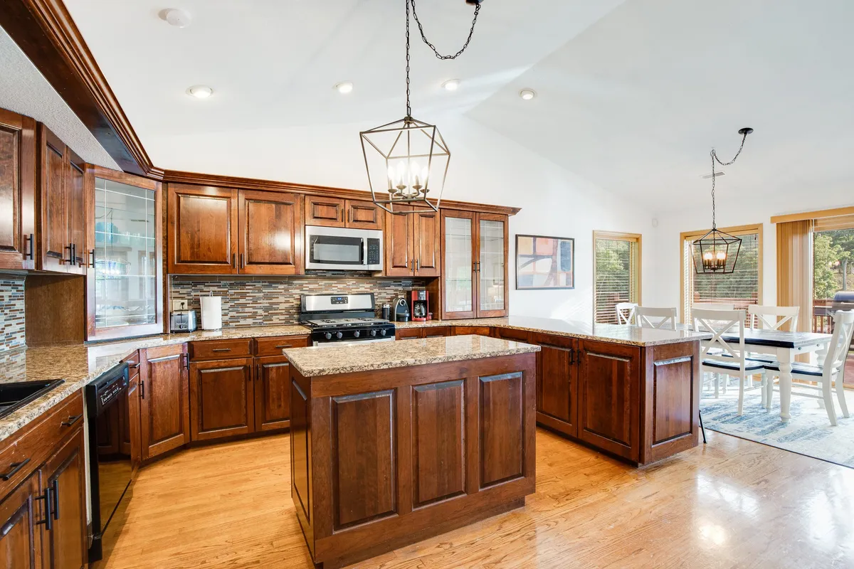 This warm, inviting kitchen and dining space showcases a granite countertop, wood cabinetry, hanging light fixtures, and a dining area with large windows. The arrangement is perfect for shared meals, celebrations, and morning coffee rituals, with direct views outside, reinforcing the open-concept design.