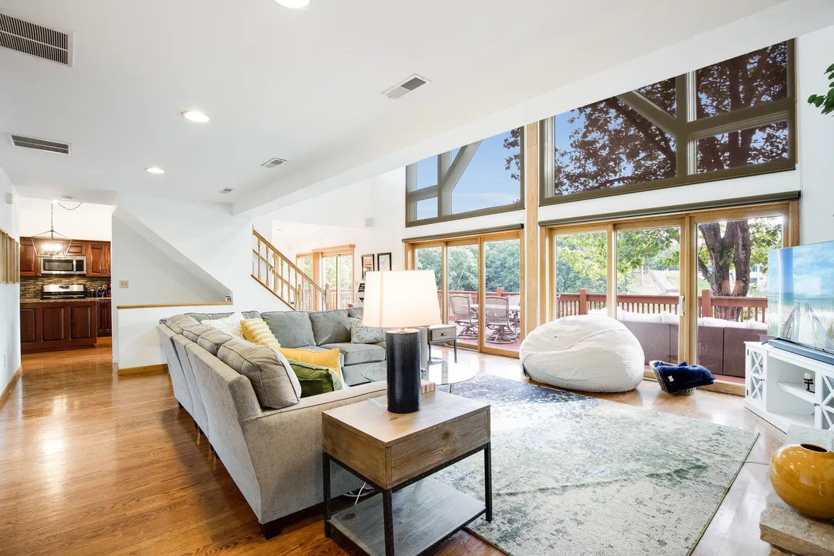 This image features a sunlit living room with large windows that flood the space with natural light. The comfortable sofa, light-colored rug, and modern coffee table create a welcoming environment for relaxing, entertaining, or gathering with friends and family. The lake views visible through the windows add a scenic backdrop and connect the interior to the outdoors.