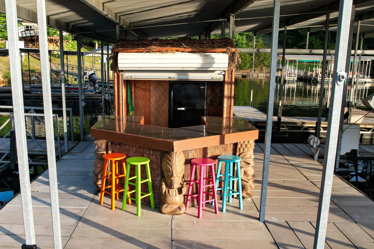 A festive interior shot of the tiki-style dock bar with colorful stools lined up, offering a direct water view just beyond. The relaxed setup invites guests to enjoy drinks and casual conversation right by the lake, with the water visible through the windows.
