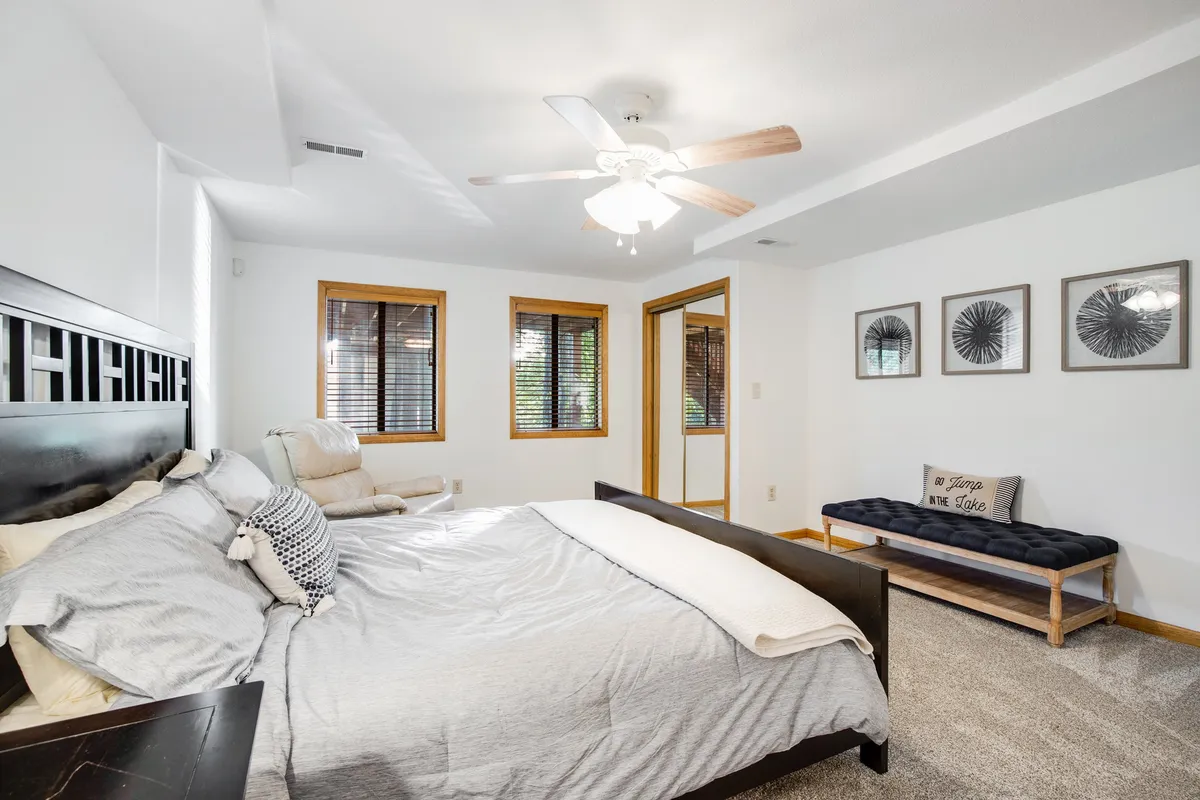 A serene bedroom with a large bed topped with decorative pillows, a plush seating area by the window, framed wall art, and abundant natural light streaming in. The space feels private and perfect for unwinding with a book or enjoying a peaceful moment alone.