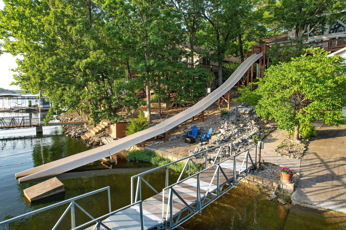 The hero image showcases a spacious lakeside home surrounded by lush trees, with a prominent two-story water slide leading straight into the lake, a private dock perfect for boating or relaxing, and inviting seating areas for family gatherings by the water.