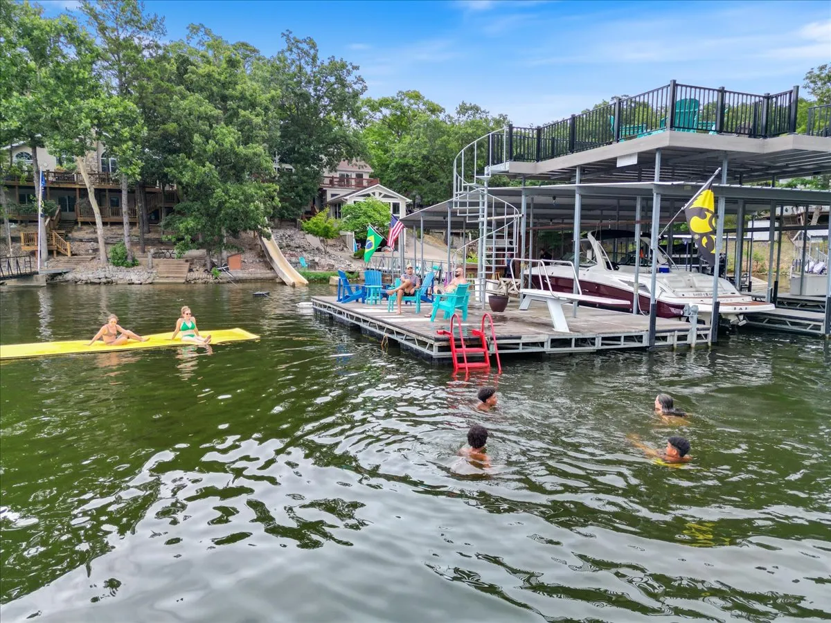 This lively exterior shot features the dock equipped with colorful chairs, children swimming, a visible water slide, a boat, and a yellow float. The scene is filled with social activity, highlighting sunbathing, boating, and relaxed lakeside fun for all ages.